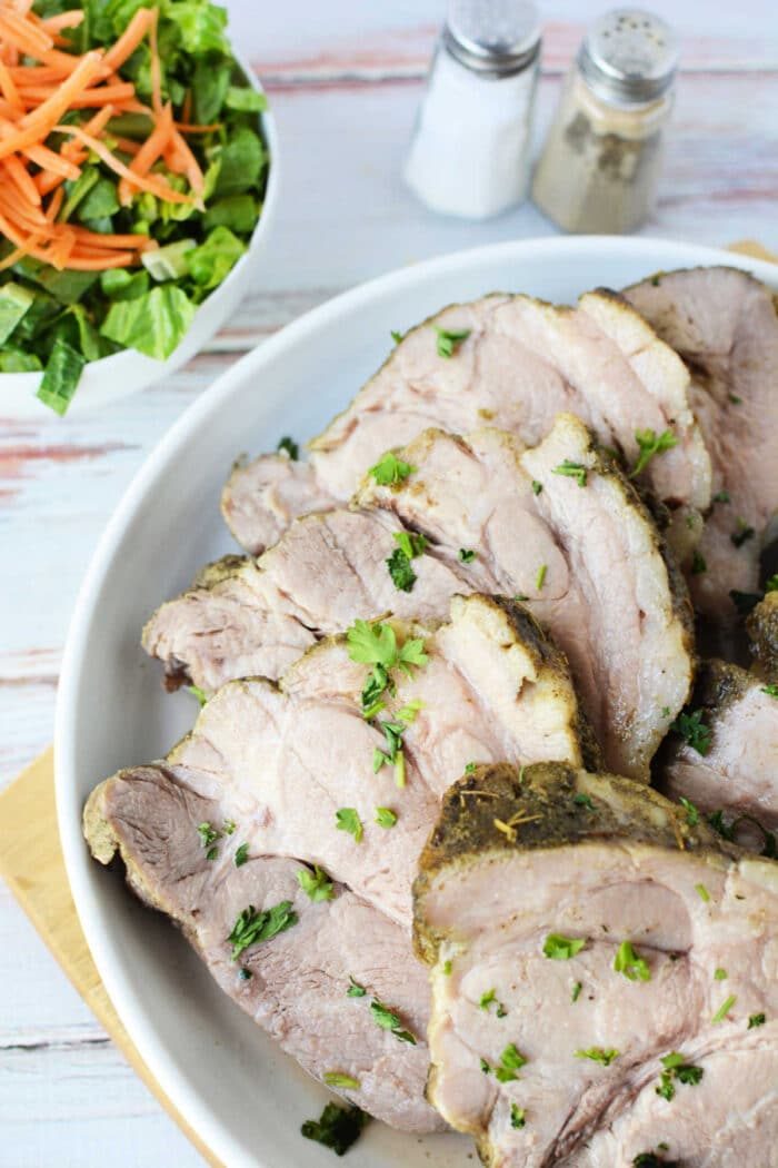 Sliced roast pork, tenderly prepared in an Instant Pot and garnished with fresh herbs, is elegantly arranged on a plate. In the background, a crisp salad accompanies the meal, alongside salt and pepper shakers.