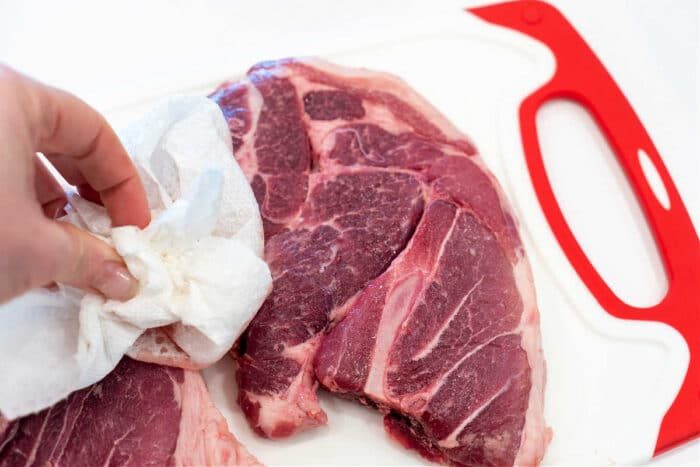 A hand dabbing raw pork shoulder steaks with a paper towel on a white cutting board with a red handle.