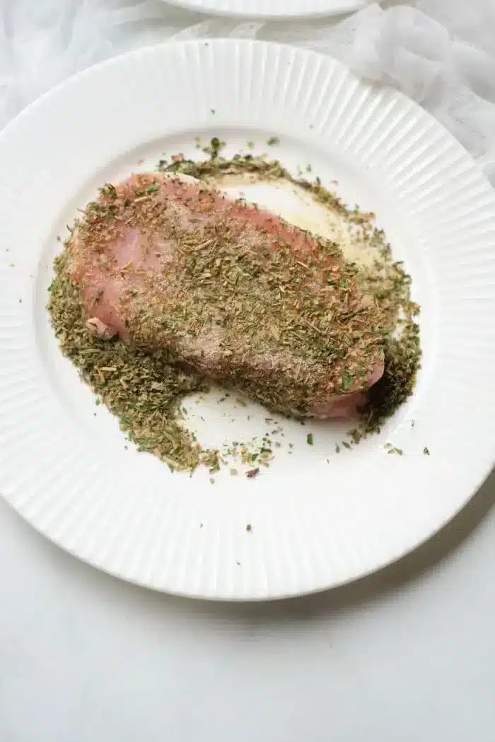 A pork steak, seasoned with a variety of herbs, rests on a white, ridged plate. The finely chopped herbs blanket the top of the meat and spill over onto the plate. solid for an air fryer, the background sets a light, textured scene.