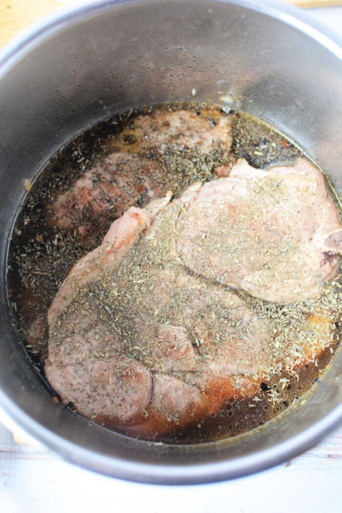 Two raw pork steaks with herbs and broth in a metal pot, ready for cooking—a simple start to a good pork steak recipe.