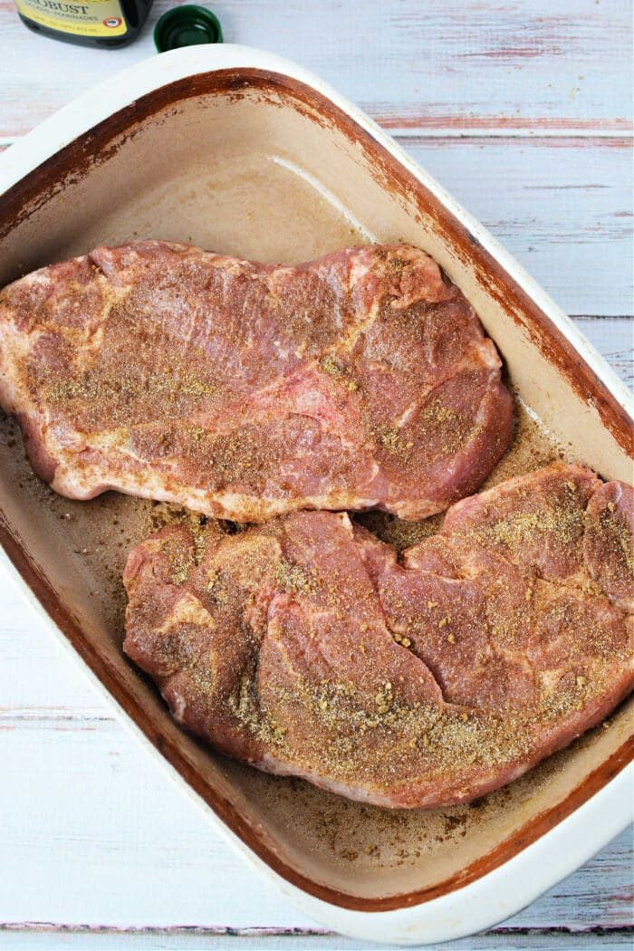 Two seasoned pork steaks in a ceramic-coated baking dish, with an olive oil bottle and wooden surface in the background, ready for roasting.