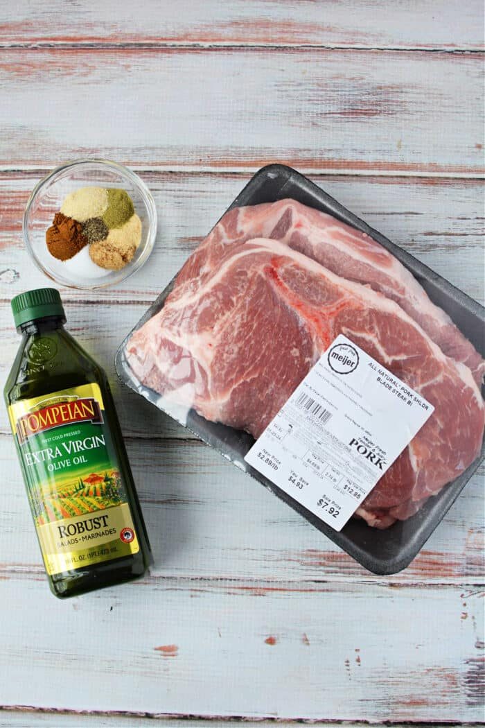 Packaged pork steaks, solid for transforming into succulent pork steaks in the oven, sit alongside a bowl of mixed spices and a bottle of olive oil on a light wooden surface.