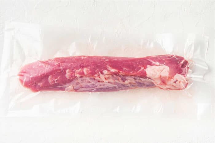 A vacuum-sealed package containing a raw, whole pork tenderloin on a plain, light-colored surface. The meat is pink with visible marbling and fat along its side—solid for making mouthwatering Pork Tenderloin Tacos.