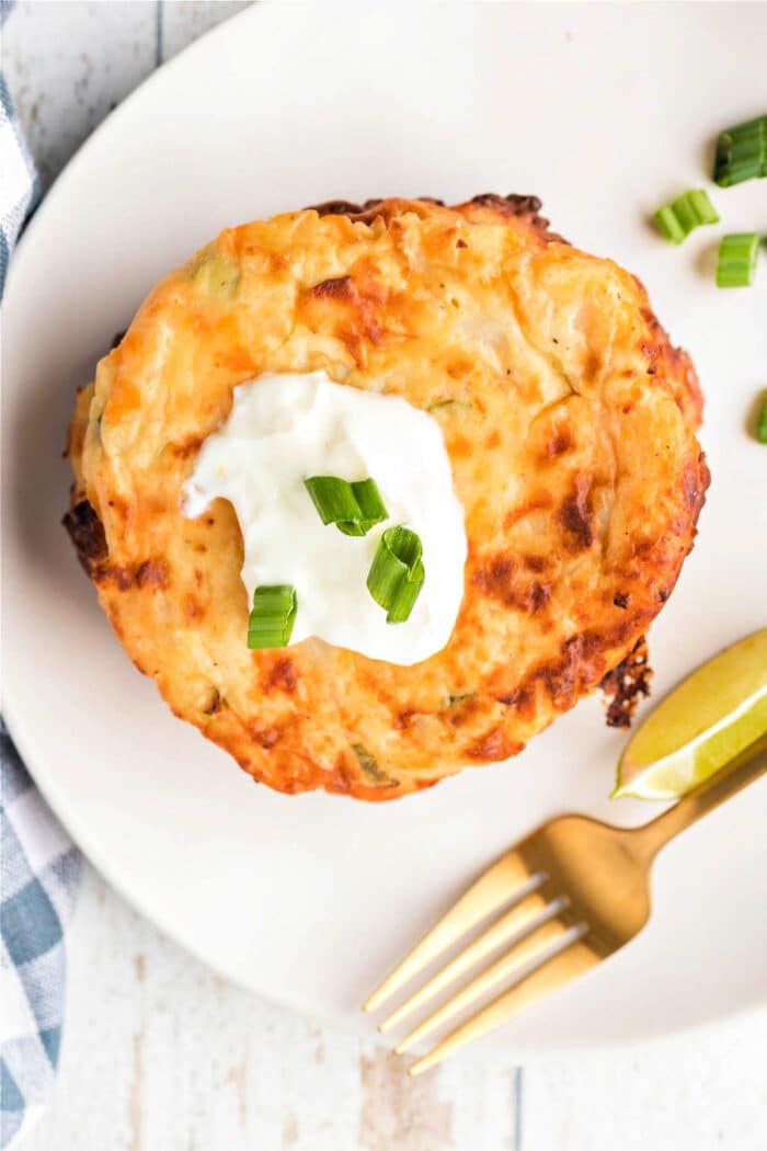 A cheesy potato pancake topped with sour cream and chopped green onions, served with lime wedges on a white plate, reminiscent of fried mashed potatoes with cheese.