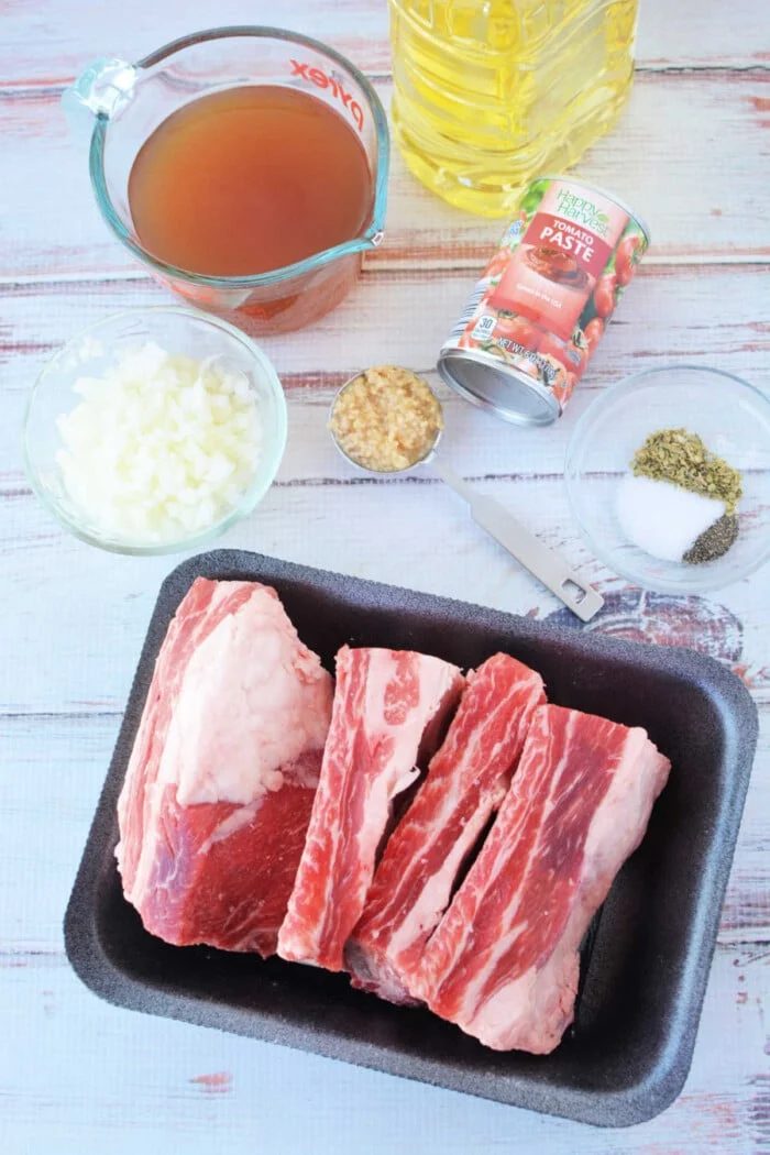 Ingredients for cooking: beef ribs, chopped onions, broth, tomato paste, minced garlic, spices, and oil on a table—solid for a savory pressure cooker recipe.