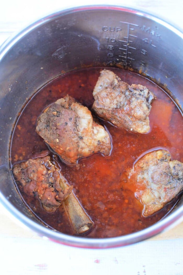Cooked oxtail in a pot with rich, red broth, hinting at the tenderness achieved by using a pressure cooker. Four pieces are visible alongside aromatic spices and herbs.