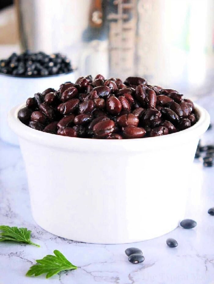 A white bowl filled with perfectly tender black beans cooked in a pressure cooker rests on a marble surface, with dried beans scattered in the background.