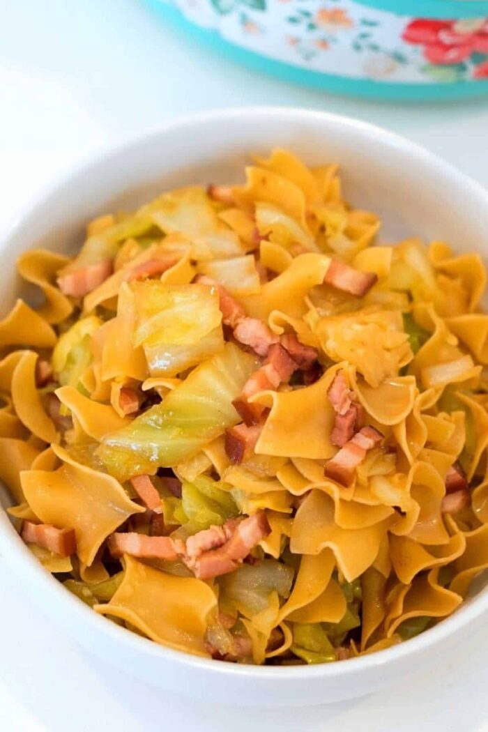 A bowl of egg noodles, infused with pressure cooker cabbage and pieces of cooked bacon. The noodles are golden, and the cabbage is lightly sautéed. This comforting dish is served in a white bowl resting on a light-colored surface.