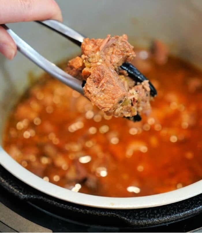 A close-up shows a hand holding metal tongs with a piece of succulent carnitas above a pot brimming with meat in a rich, red sauce. The pot appears to be part of a pressure cooker, ensuring the flavors meld perfectly.