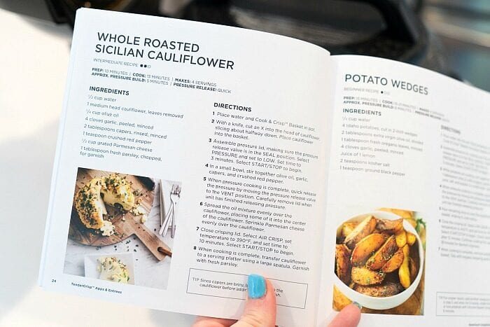A hand holds open a cookbook, displaying recipes for roasted cauliflower—including whole pressure cooker cauliflower—and potato wedges alongside vibrant recipe photos.