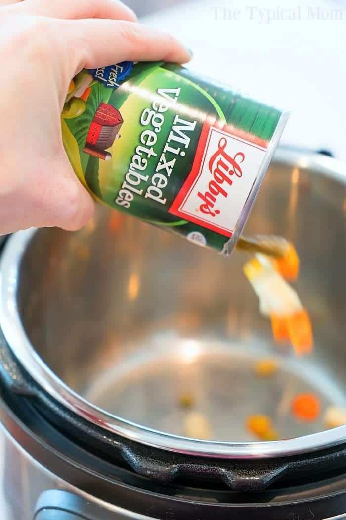 Hand pouring canned mixed vegetables into a pressure cooker for a cozy chicken pot pie soup.