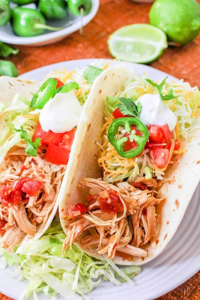 Two Pressure Cooker Chicken Tacos with lettuce, cheese, tomatoes, jalapeños, and sour cream served on a white plate.