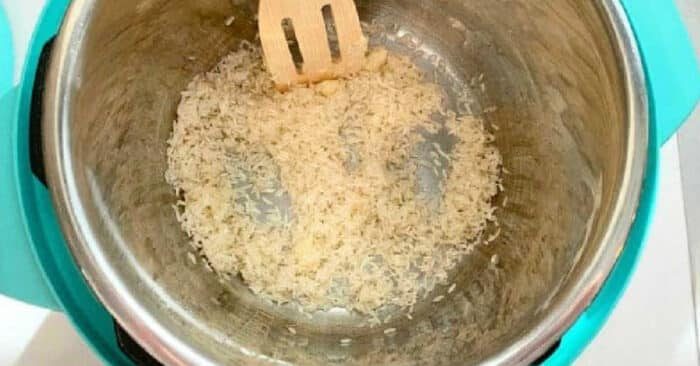 Uncooked rice being stirred with a wooden spatula in a stainless steel pot on a teal base, ready to become a good pressure cooker coconut rice pudding.