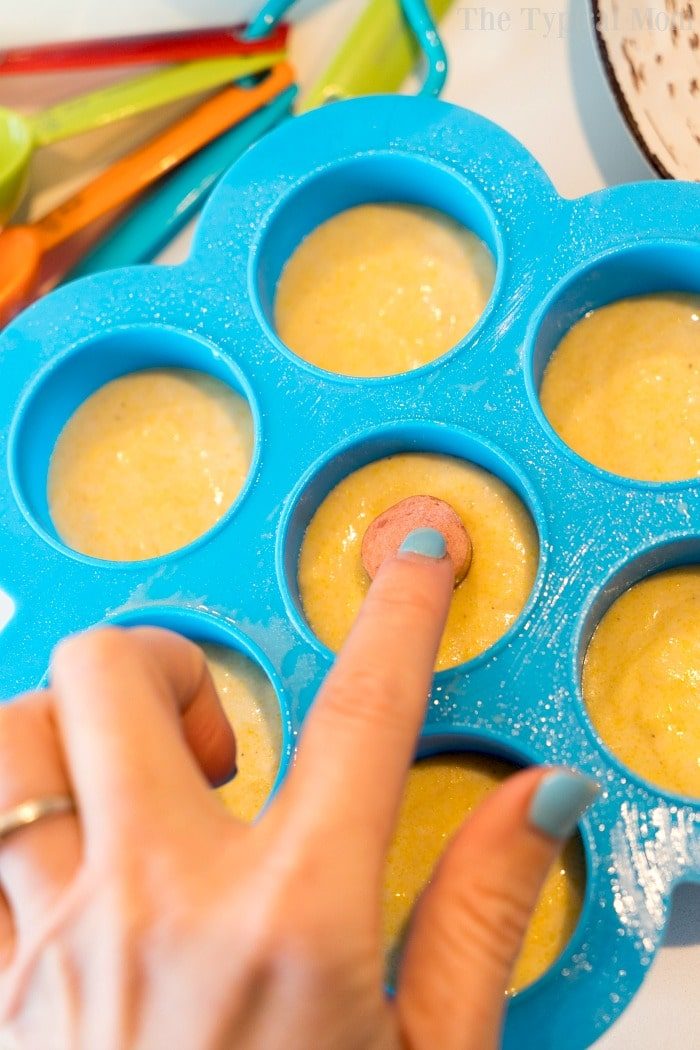 A hand places a mini hot dog into cornbread batter in a blue silicone mold, preparing pressure cooker corn dog bites. Colorful measuring spoons are nearby.