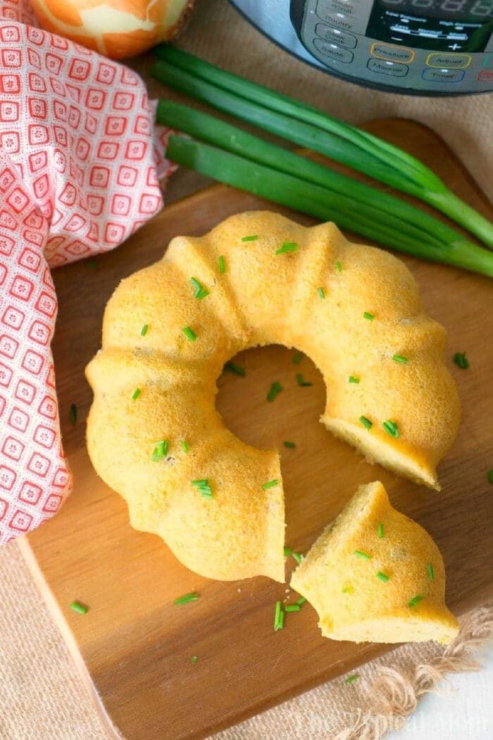 A sliced pressure cooker cornbread bundt garnished with chives sits on a wooden board, next to green onions and a red towel.