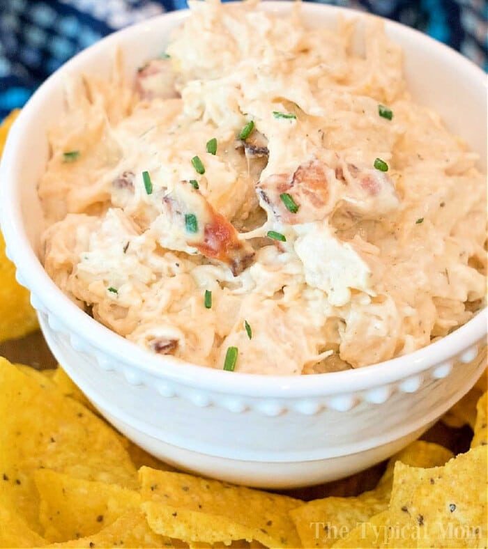 A white bowl brimming with creamy pressure cooker crack chicken dip, garnished with chopped chives. The bowl is surrounded by yellow tortilla chips on a table, inviting you to dive into this good creation.