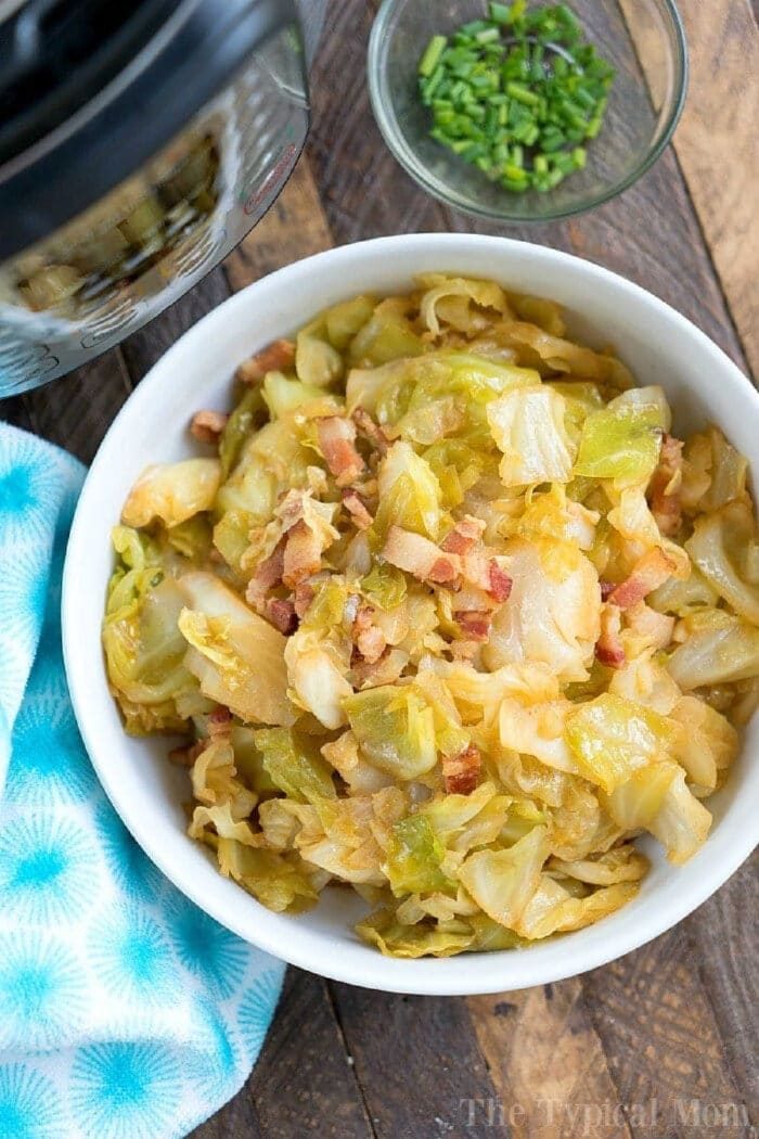 Bowl of pressure cooker fried cabbage with bacon pieces, next to a small bowl of chopped chives on a wooden table.