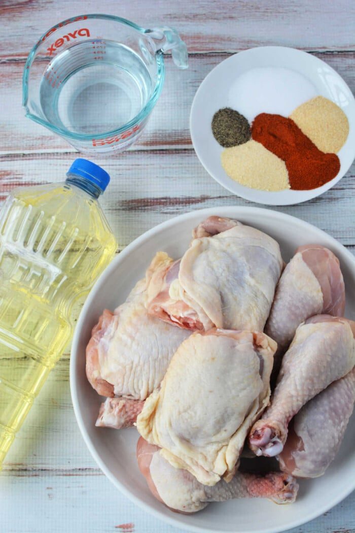 A raw chicken awaits transformation beside an oil bottle and a measuring cup of water, with a plate of spices ready to spice things up. solid ingredients for pressure cooker magic or crispy fried chicken bliss, all resting on a light wood surface.