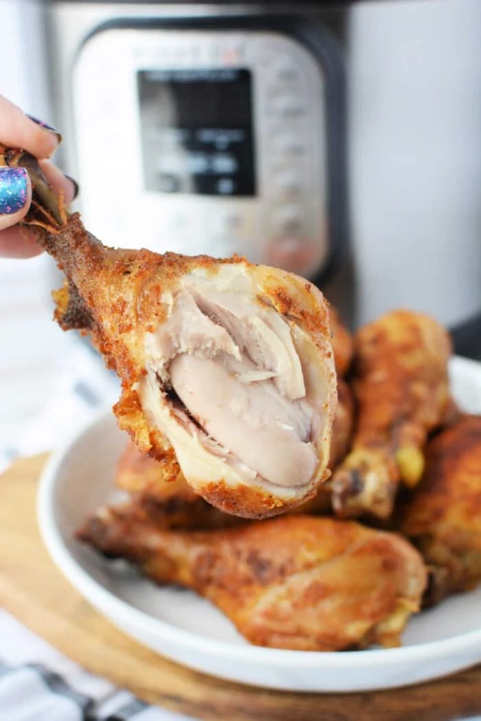 Close-up of a hand holding a juicy piece of pressure cooker fried chicken with visible meat, set against the backdrop of a gleaming pressure cooker.