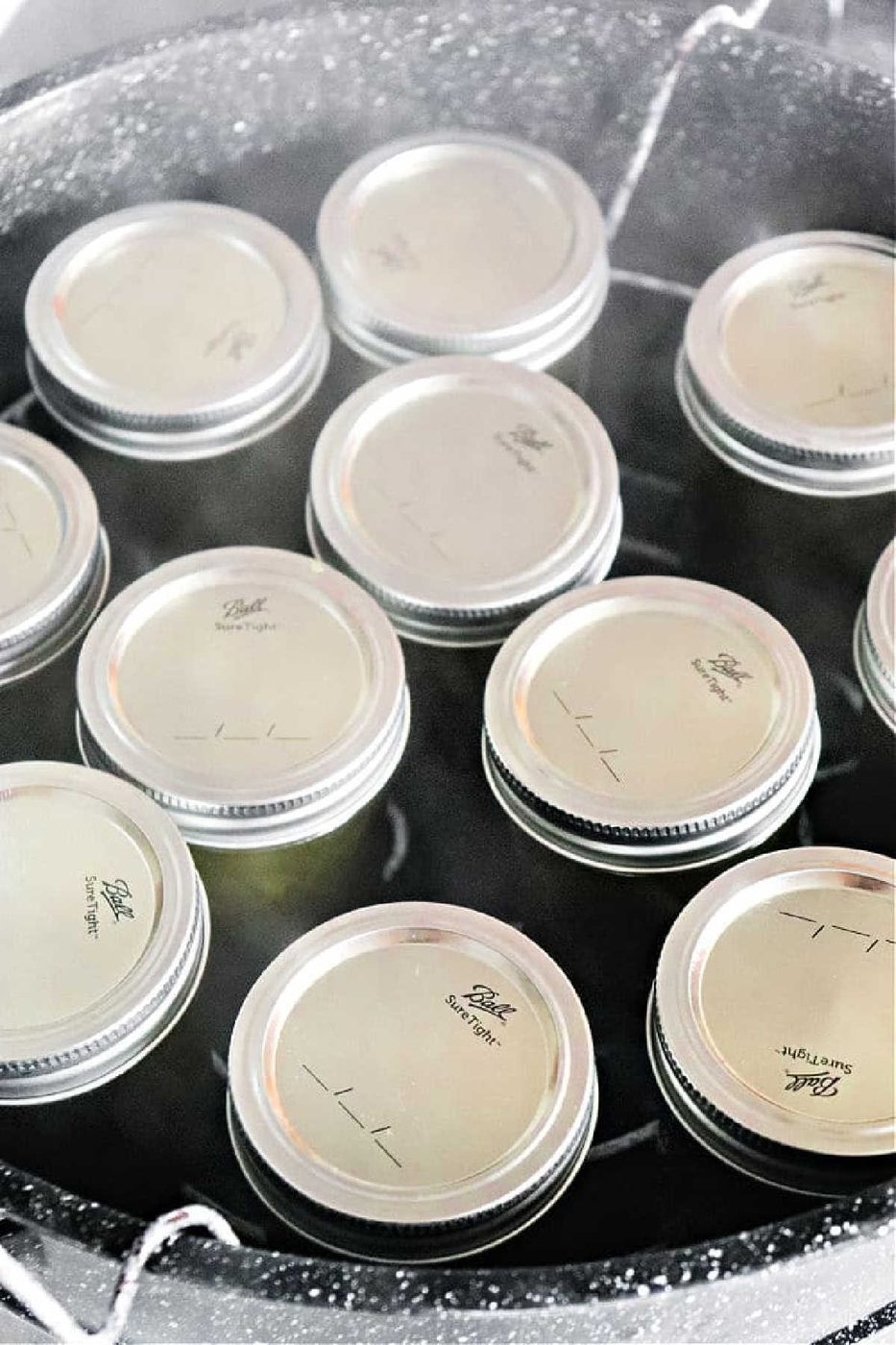 Twelve sealed canning jars are being processed in a large pot, similar to how you'd make pressure cooker jam, with hot water surrounding each jar.