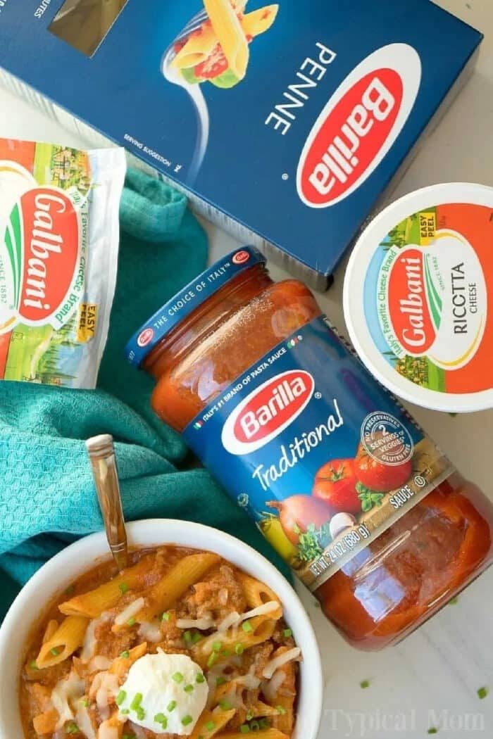 Penne pasta, ricotta cheese, and pasta sauce surround a bowl of pressure cooker lasagna casserole, topped with cheese and fresh herbs.