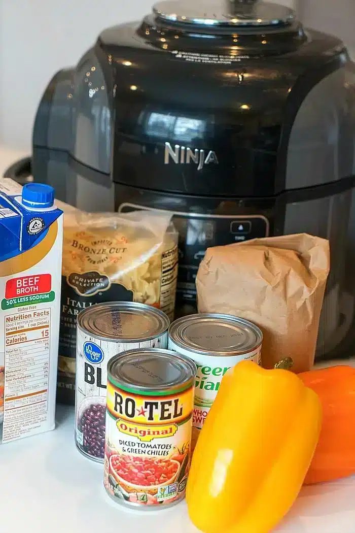 An assortment of kitchen items in front of a black Ninja pressure cooker: beef broth carton, bag of pasta, brown paper bag, canned black beans, diced tomatoes with green chilies, canned peas, and a yellow bell pepper—solid for whipping up a spicy Mexican casserole.