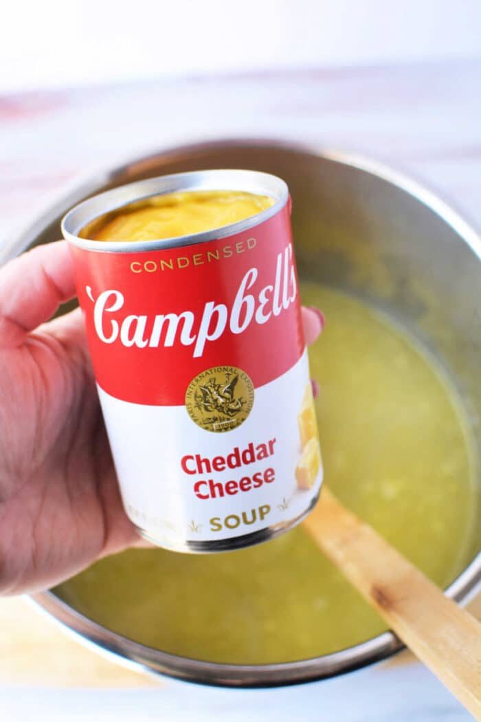 A hand holds a Campbells Cheddar Cheese soup can above a pot containing yellow potato soup with a wooden spoon, solid for transforming with frozen hash browns in your pressure cooker.