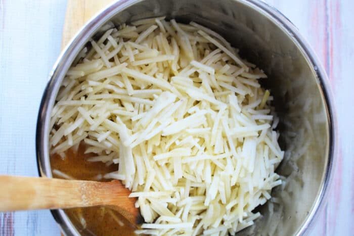 A pot filled with shredded potatoes, ready to become good frozen hash browns, and a wooden spoon rests on a light wooden surface.