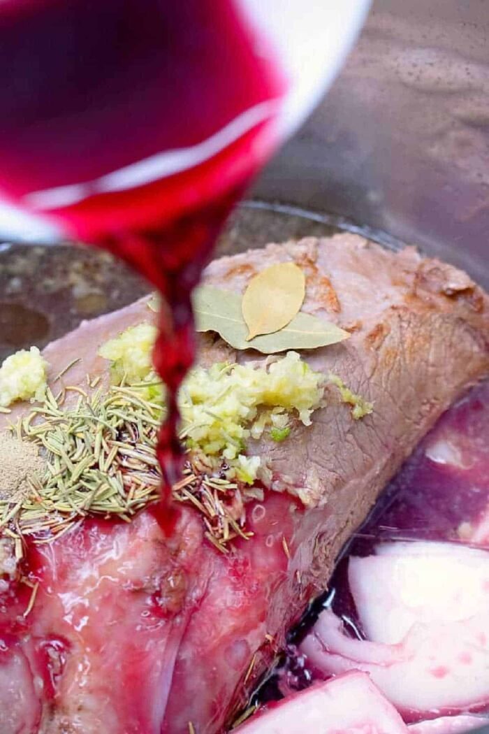 Red wine being poured over a pressure cooker roast beef seasoned with herbs, garlic, and a bay leaf.