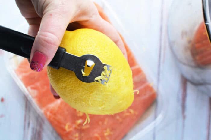 A hand expertly uses a zester to zest a lemon over a fresh container of salmon, ready to be transformed into a delightful dish. Consider using your pressure cooker for perfectly tender and flavorful results in no time.
