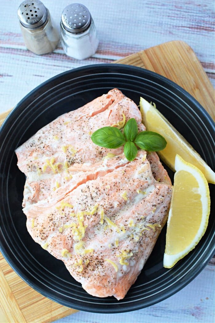 Salmon fillets with lemon zest and basil on a black plate, served alongside lemon wedges, salt, and pepper shakers. Perfectly tender, these fillets could easily be mistaken for being made in a pressure cooker.
