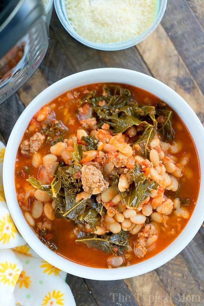A bowl of hearty Italian sausage soup with beans and kale, prepared effortlessly in a pressure cooker, sits on a wooden table next to freshly grated cheese.