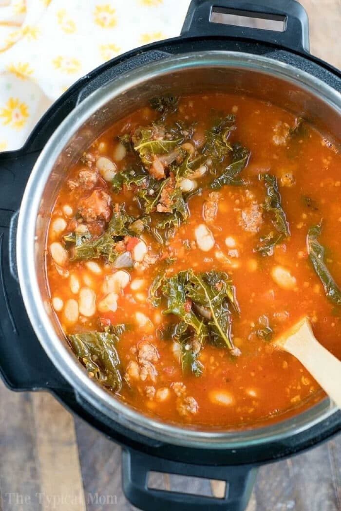A pot of pressure cooker sausage soup with beans, leafy greens, and tomato broth is being stirred with a wooden spoon.