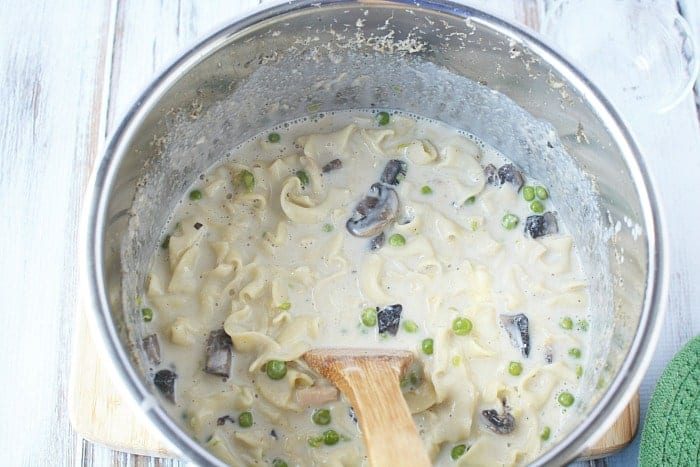 A pot of creamy tuna noodle pasta with mushrooms and peas, reminiscent of a classic pressure cooker tuna noodle casserole, is gently stirred with a wooden spoon on a rustic wooden surface.