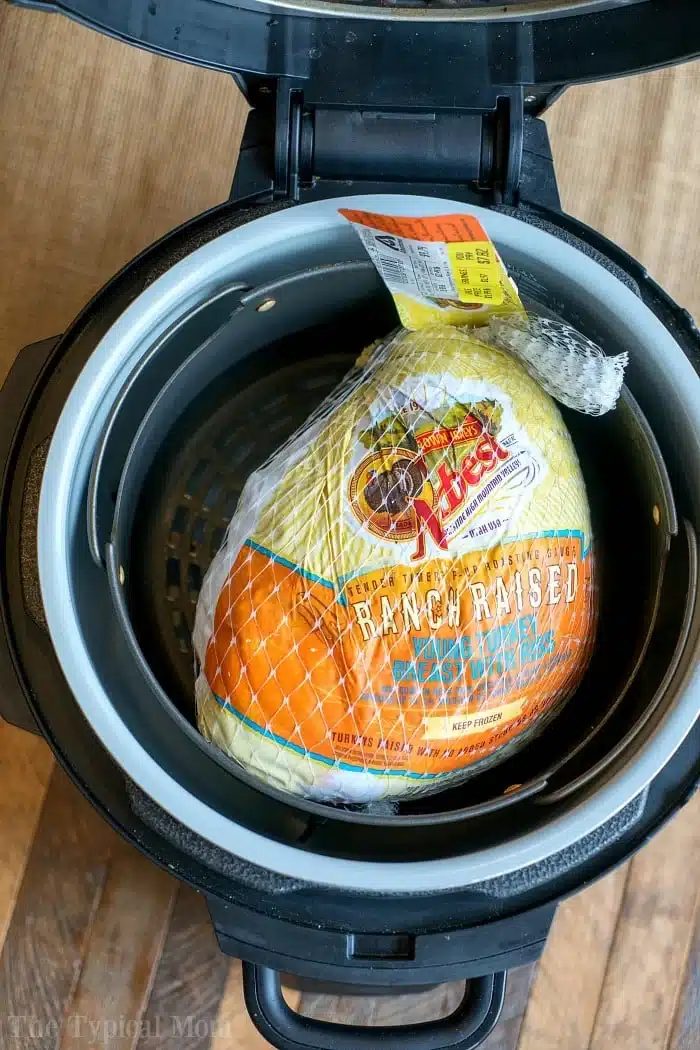A frozen whole turkey in packaging sits inside an open Instant Pot pressure cooker, ready to transform into a juicy pressure cooker turkey breast on a wooden surface.