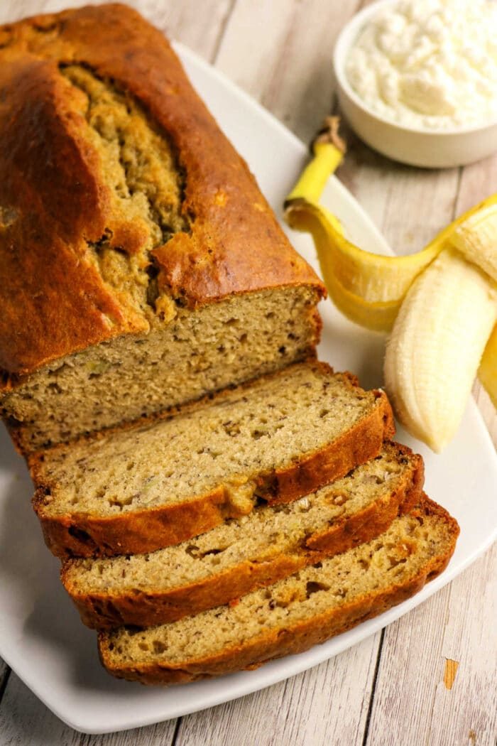 Sliced Protein Banana Bread on a plate with a peeled banana and a bowl of cottage cheese in the background.