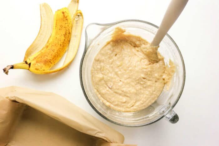A glass bowl of Protein Banana Bread batter with a spatula sits beside banana peels and a parchment-lined pan, ready to be baked.