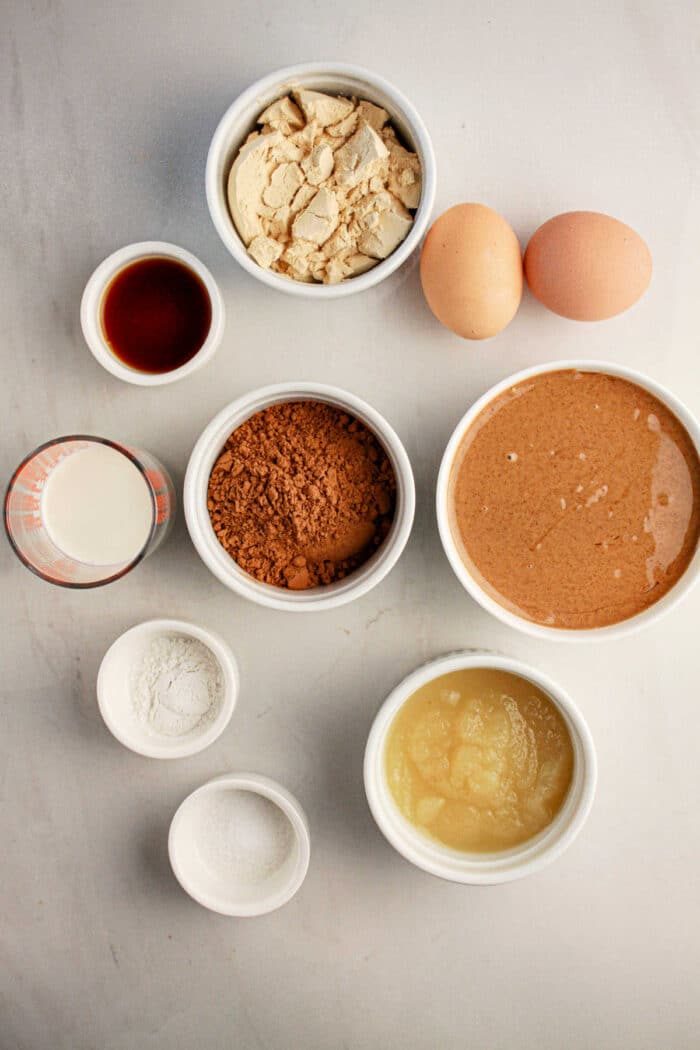 Ingredients for a protein brownie recipe on the table: two eggs, vanilla, coconut flour, cocoa, peanut butter, milk, applesauce, baking powder, and salt.