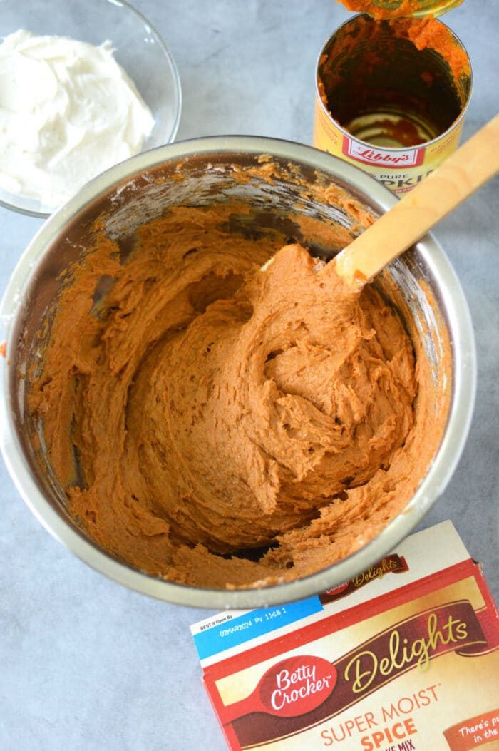 A mixing bowl filled with pumpkin cake bars batter sits on a gray surface, surrounded by canned pumpkin and a cake mix box, ready to whip up good pumpkin bars with cake mix. Nearby, whipped topping waits to add the finishing touch.