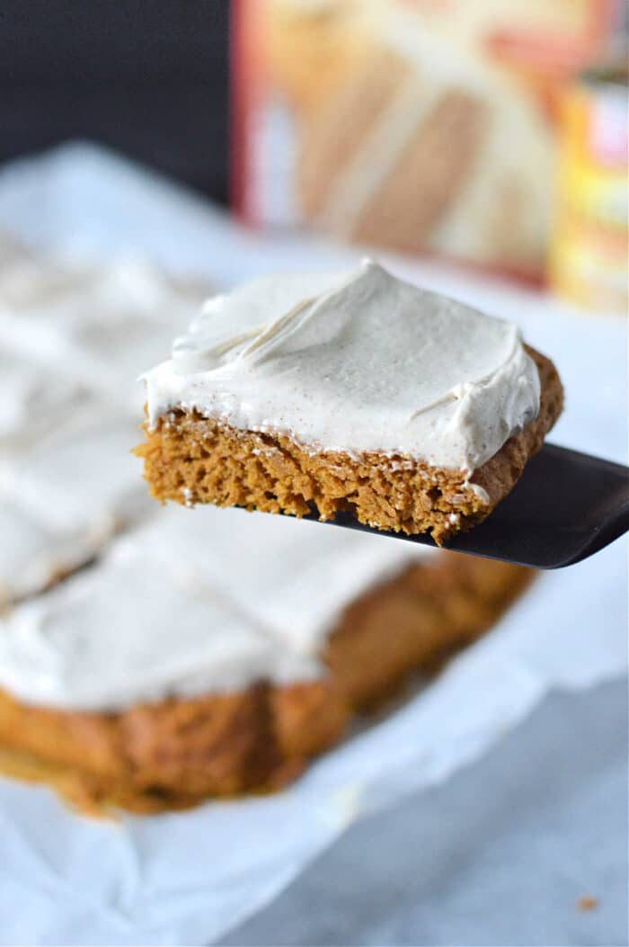 A frosted square pumpkin bars with cake mix, is lifted on a spatula above more slices on parchment paper.