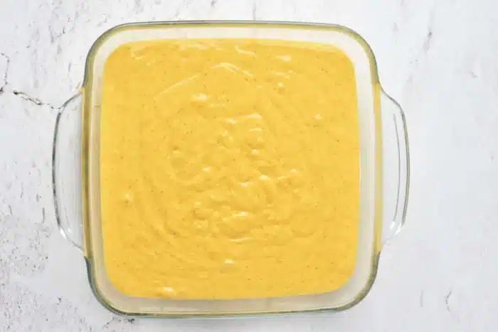 A square glass dish filled with a smooth, yellow batter sits on a light-colored marbled surface. The batter, solid for pumpkin cake with yellow cake mix, has a few small bubbles scattered across the top, indicating that it has been prepared and is ready to be baked.