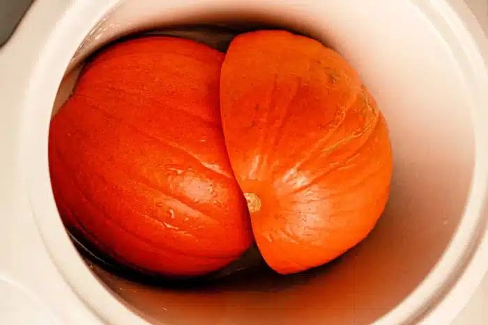 A Pumpkin in a Slow Cooker, cut in half with the two halves placed next to each other. The bright orange flesh of the pumpkin is visible, and there is slight moisture on the surface. The cooker has a white interior.