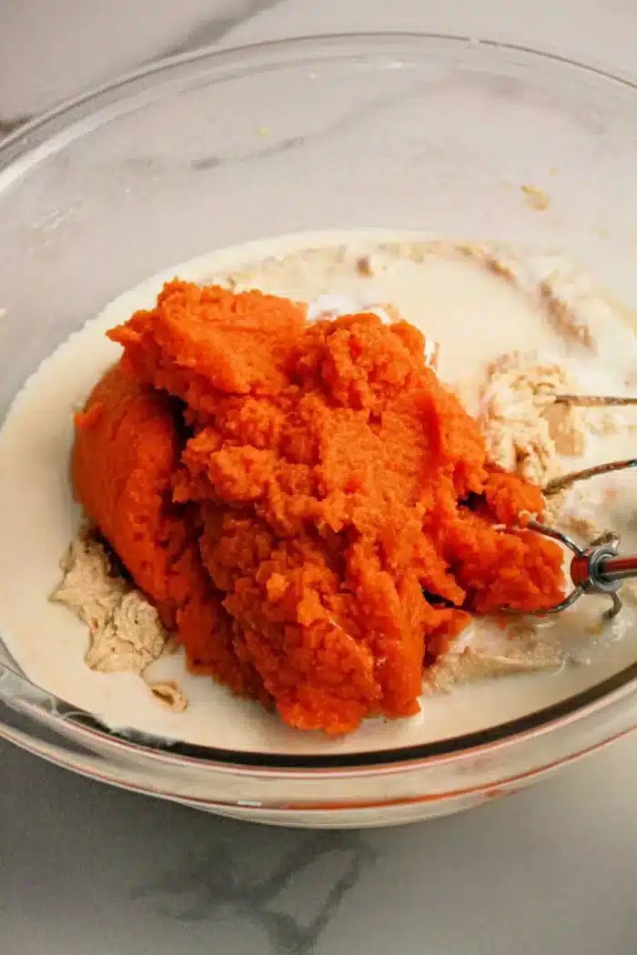 A glass mixing bowl containing a vibrant orange pumpkin puree, pale beige dry ingredients, and a liquid. An electric mixer beater partially submerged in the mixture is visible on the right side of the bowl, ready to transform these elements into a good Pumpkin Poke Cake.