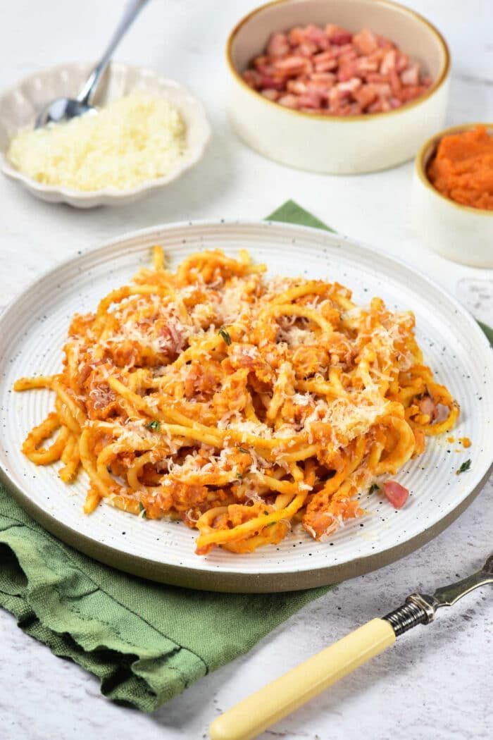 Plate of pasta with red sauce and grated cheese, featuring a good twist of Pumpkin Spaghetti, with small bowls of cheese, diced ham, and sauce in the background.