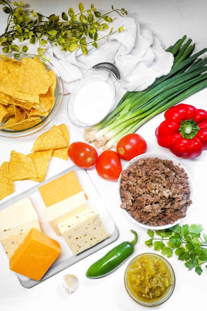 Taco dip ingredients: chips, cheese blocks, ground beef for a hearty Queso Dip with Meat, fresh tomatoes, green onions, pepper, milk, and seasonings.