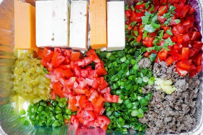 Chopped vegetables, ground meat, and cheese blocks arranged in sections in a foil tray—solid for assembling a Queso Dip with Meat.