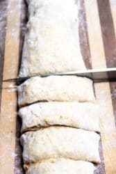 A knife slices through a cylindrical piece of dough, destined to become cinnamon roll muffins, on a wooden surface. The lightly floured dough and the flour-dusted surface hint at the sweet transformation awaiting these evenly cut sections.