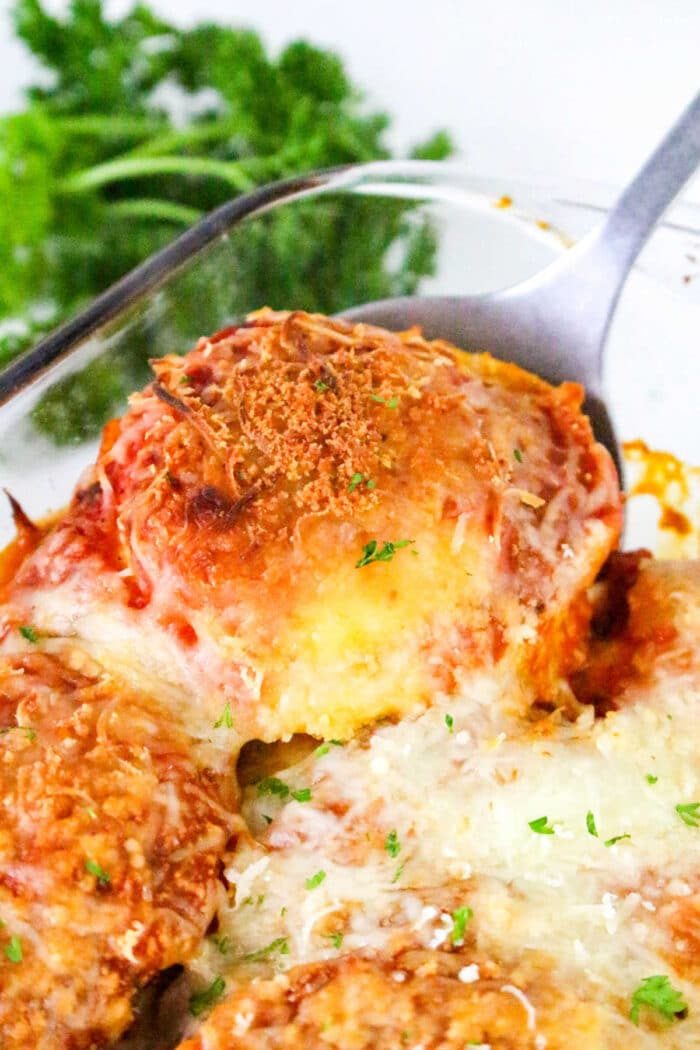 A serving of cheesy, baked eggplant parmesan is lifted from a glass dish, garnished with herbs—solid alongside a comforting Ravioli Casserole.