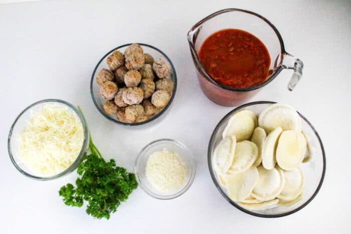 Bowls of shredded cheese, frozen meatballs, tomato sauce, ravioli, grated cheese, and fresh parsley on a white surface—everything you need to create a good Ravioli Casserole.