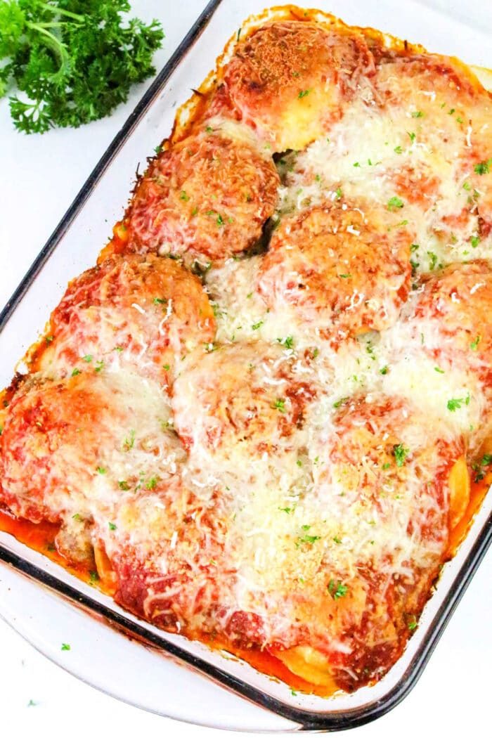 Baked chicken parmesan in a casserole dish, topped with melted cheese and parsley, served alongside a hearty ravioli casserole for a comforting meal.